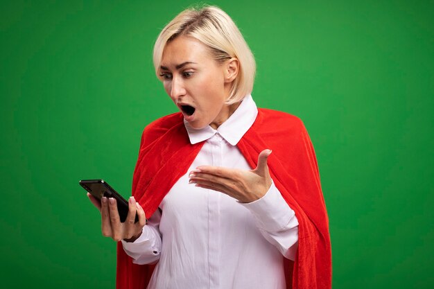 Surprised middle-aged blonde superhero woman in red cape holding and looking at mobile phone pointing at it with hand isolated on green wall with copy space