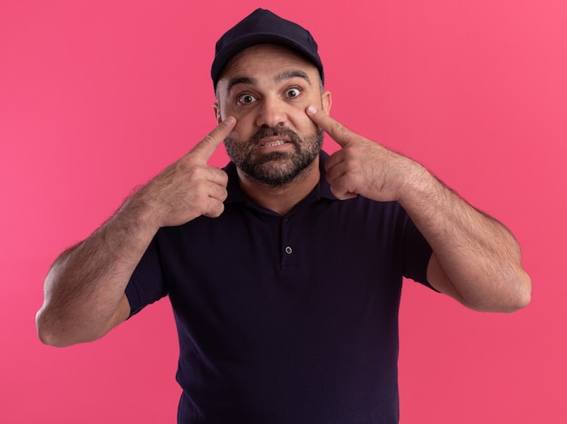 Surprised middle-aged delivery man in uniform and cap pulling down eye lids with fingers isolated on pink wall