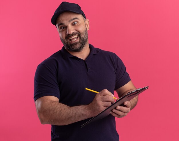 Surprised middle-aged delivery man in uniform and cap writing something on clipboard isolated on pink wall