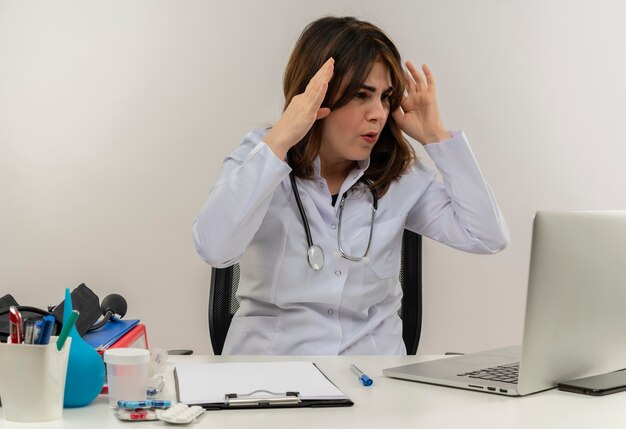 Surprised middle-aged female doctor wearing medical robe and stethoscope sitting at desk with medical tools clipboard looking at laptop touching head isolated