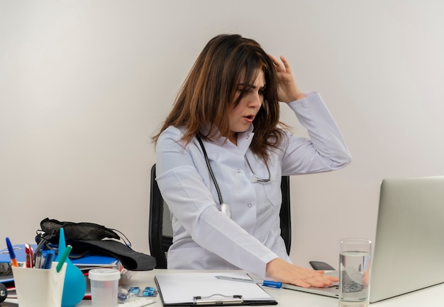 Free photo surprised middle-aged female doctor wearing medical robe and stethoscope sitting at desk with medical tools clipboard using and looking at laptop touching head isolated