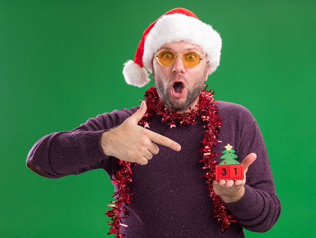 Free Photo surprised middle-aged man wearing santa hat and tinsel garland around neck with glasses holding and pointing at christmas tree toy with date looking at camera isolated on green background