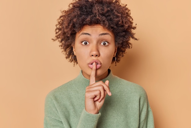 Surprised mysterious woman makes silence gesture presses index finger over lips tells gossips keeps eyes widely opened dressed in casual jumper isolated over brown background. Hush don not speak