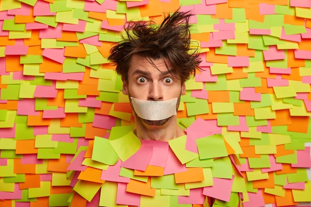 Free photo surprised nervous man with adhesive duct tape over mouth, asks be silent, stands silenced and speechless, poses against colorful wall with sticky notes, being frightened. shut up, censorship