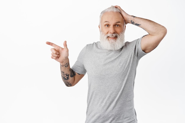 Surprised old guy pointing left and touching head, smiling amazed, encounter something interesting, have you seen it gesture, look amazed at camera, standing over white background.