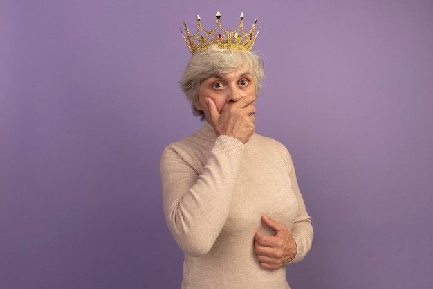 Free photo surprised old woman wearing creamy turtleneck sweater and crown putting hand on mouth and on belly