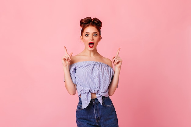 Free photo surprised pinup girl pointing up with fingers. front view of shocked ginger lady in blouse isolated on pink space.