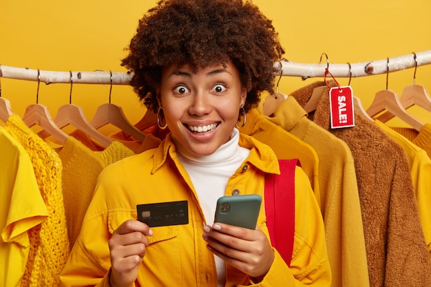 Surprised positive African American woman smiles broadly, uses modern smartphone and credit card