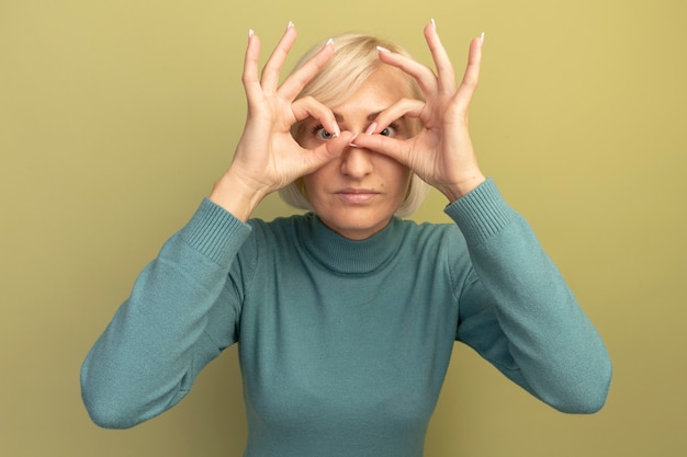 Free Photo surprised pretty blonde slavic woman looks at camera through fingers on olive green