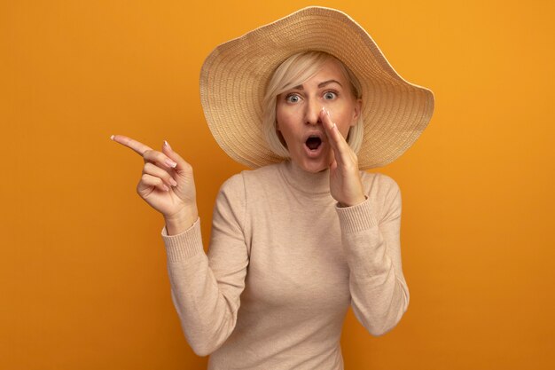 Surprised pretty blonde slavic woman with beach hat holds hand close to mouth and points at side on orange