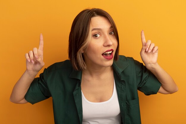 Surprised pretty caucasian woman points up with two hands on orange