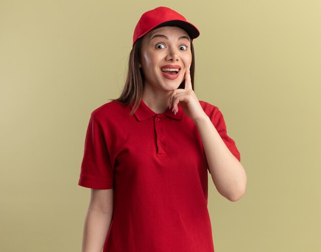 Surprised pretty delivery woman in uniform puts finger on face and looks at camera