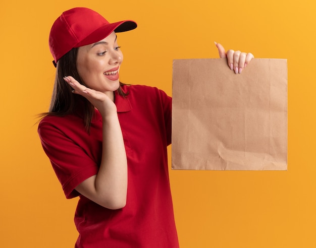 Surprised pretty delivery woman in uniform stands with raised hand holding and looking at paper package isolated on orange wall with copy space