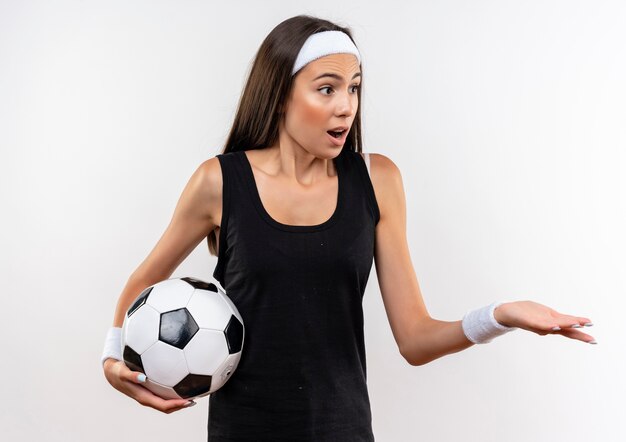 Surprised pretty sporty girl wearing headband and wristband holding soccer ball looking at side and showing empty hand isolated on white space