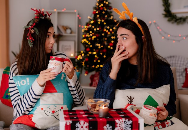 surprised pretty young girls with holly wreath and reindeer headband hold cups looking at each other sitting on armchairs and enjoying christmas time at home