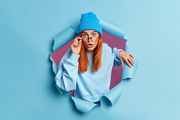 Surprised redhead European woman has shocked face expression stares with eyes popped out reacts on astounding news wears casual jumper and hat.