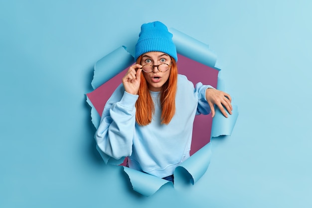 Surprised redhead European woman has shocked face expression stares with eyes popped out reacts on astounding news wears casual jumper and hat.