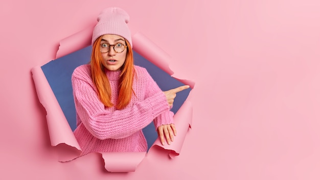 surprised redhead woman feels astonished, pointing at copy space, wears knitted hat and sweater