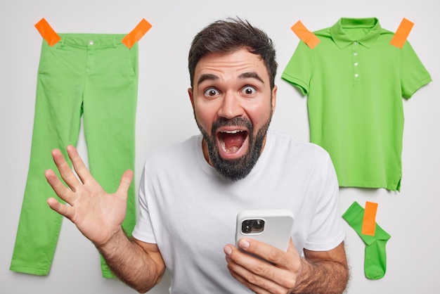 Free Photo surprised shocked bearded young man holds mobile phone reacts on amazing news exclaims loudly dressed in casual basic t shirt poses indoor with green clothes hanging behind sells clothing online