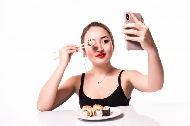 Surprised smiling asian woman cover her eye with sushi roll and make selfie on her phone