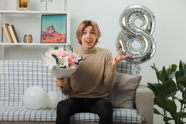 Surprised spreading hand handsome guy on happy women day holding bouquet sitting on sofa in living room