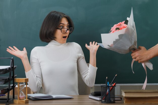 surprised spreading hands young female teacher sitting at desk with school tools received a bouquet of flowers in classroom