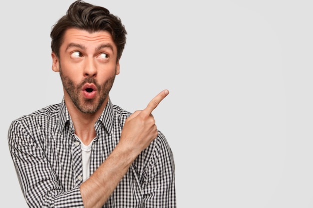 Surprised stupefied European young male with shocked expression, wears casual checkered shirt, points with index finger at upper right corner, has dark bristle, isolated over white wall