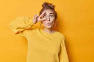 Free photo surprised teenage girl keeps lips folded makes peace gesture with fingers over eye shows salute sign wears big round silver earrings and jumper isolated over yellow background. body language concept