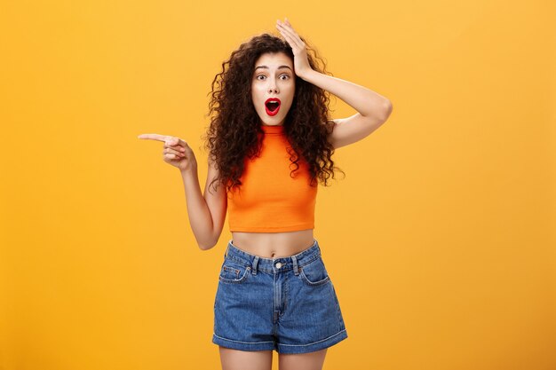 Surprised thrilled beautiful caucasian girl with curly hairstyle in red lipstic and orange cropped top holding hand on head amazed and pointing left questioned and astonished over studio background.