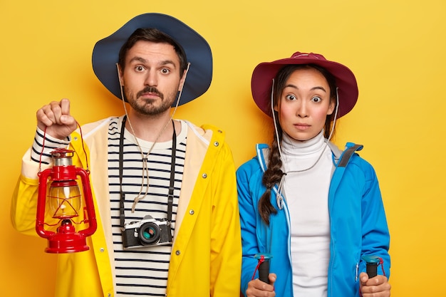 Free Photo surprised tourist couple have recreation time in nature, have journey on foot, stand with trekking sticks, oil lamp for lightening in darkness stand closely over yellow wall