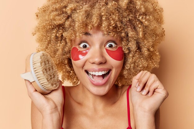 Surprised very glad young woman stares with eyes full of happiness smiles broadly holds body brush applies red moisturizing patches under eyes after taking shower poses indoor hears great news