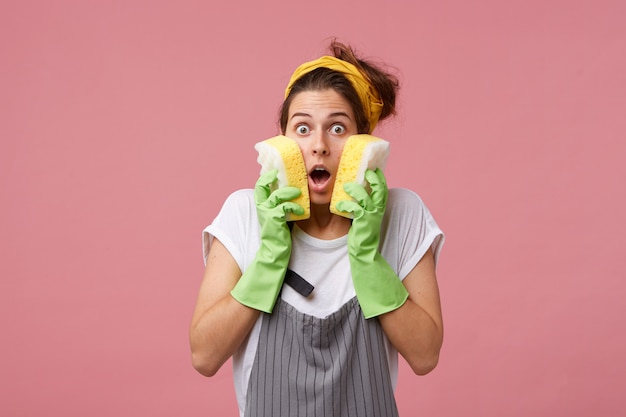 Free Photo surprised woman in apron and casual clothes wearing rubber green gloves holding two tidy sponges on cheeks realising that she should do much work. astonished female going to do her housework