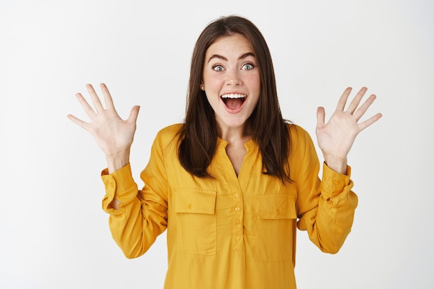 Surprised woman gasping and shaking hands amazed, receive presents and celebrating, standing over white wall.