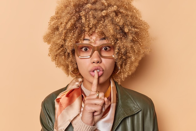 Free photo surprised woman makes taboo gesture silence sign shushes indoor looks mysterious expression dressed in stylish clothes wears eyeglasses isolated over brown background. shh keep my secret safe