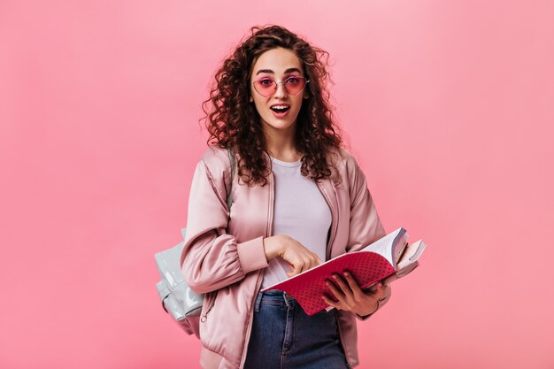 Surprised woman in pink jacket and jeans posing with books on isolated background