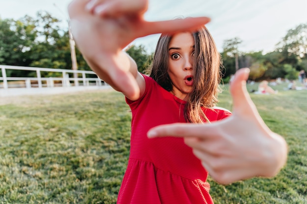 Free Photo surprised woman in red attire posing outdoor and making funny faces. photo of emotional brown-haired girl chilling in spring day.