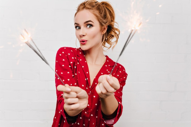 Free photo surprised woman in red pajama posing with sparkler. cute blonde lady in funny night-suit holding bengal lights in christmas morning.