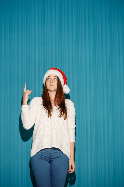 Free photo surprised woman wearing a santa hat pointing up on blue studio background