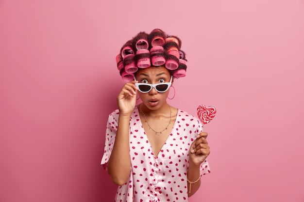 Free photo surprised woman with curlers in hair, prepares for womens day, wants to have brilliant look, wears bathrobe and sunglasses, holds delicious appetizing lollipop, isolated over pink wall