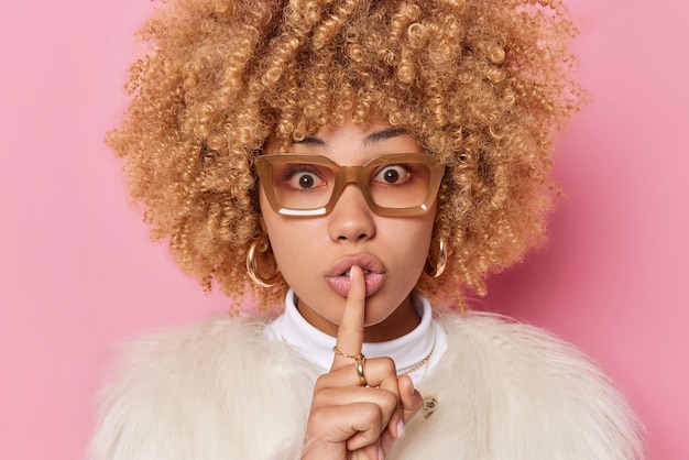 Free Photo surprised woman with curly bushy hair makes silence gesture tells secret wears spectacles and white fur coat isolated over pink background. hush do not spread rumors please. body language concept