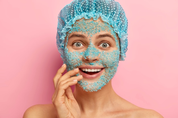 Free Photo surprised woman with happy look, covers face with blue granules of sea salt, peels from pores and acnes, wears protective headgear during taking bath