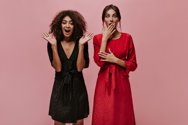 Surprised woman with round earrings in polka dor red dress looking into camera and posing with mulatto girl in black fashionable outfit