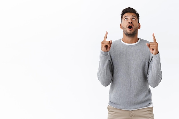 Surprised wondered handsome bearded guy with stylish haircut wear casual grey sweater drop jaw impressed raise head and staring upwards from amazement pointing up with interest and curiosity