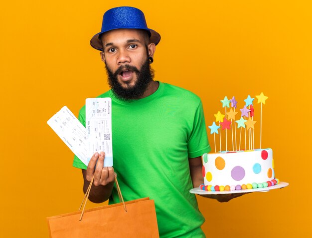 Surprised young afro-american guy wearing party hat holding gift bag and cake with tickets isolated on orange wall