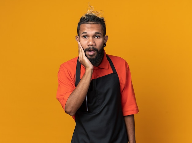 Surprised young afro-american male barber wearing uniform keeping hand on face 