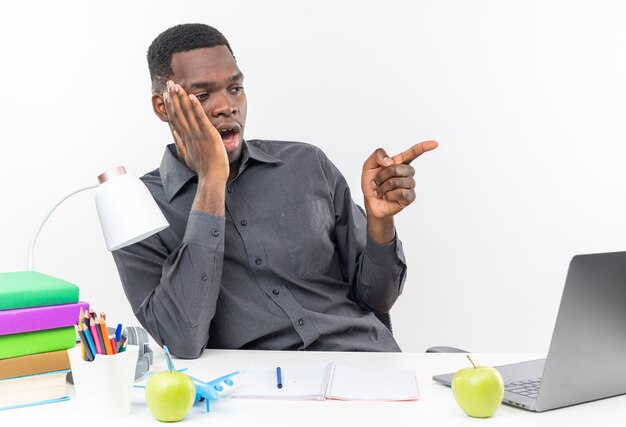 Surprised young afro-american student sitting at desk with school tools putting hand on his face looking and pointing at side 