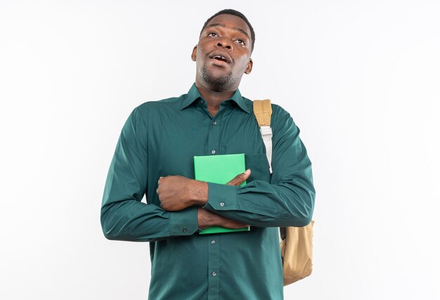 Surprised young afro-american student with backpack holding book and looking up isolated on white wall with copy space