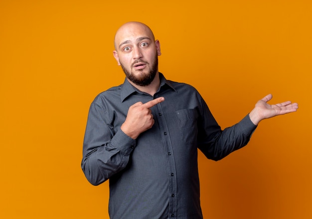 Surprised young bald call center man showing empty hand and pointing at it isolated on orange wall