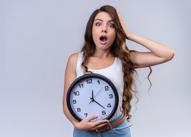 Surprised young beautiful girl holding clock and putting hand on head on isolated white wall with copy space