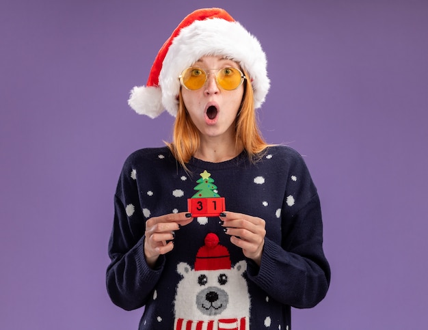 Free Photo surprised young beautiful girl wearing christmas sweater and hat with glasses holding christmas toy isolated on purple background